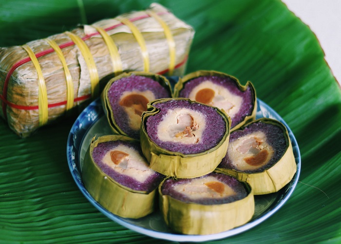 Banh Tet La Cam (Purple-Colored Sticky Rice Cake) stands out with its beautiful purple sticky rice layer created from natural purple leaves. When sliced, the cake clearly reveals the layers of yellow mung bean filling, orange salted egg yolk, and rich fatty pork in the center.